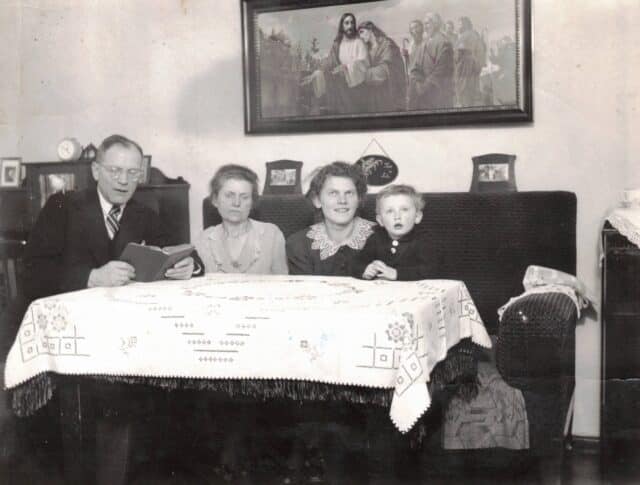 Familie Hempel sitzt mit Enkel Hans im Wohnzimmer und hört Walter Hempel zu, der aus der Bibel vorliest.