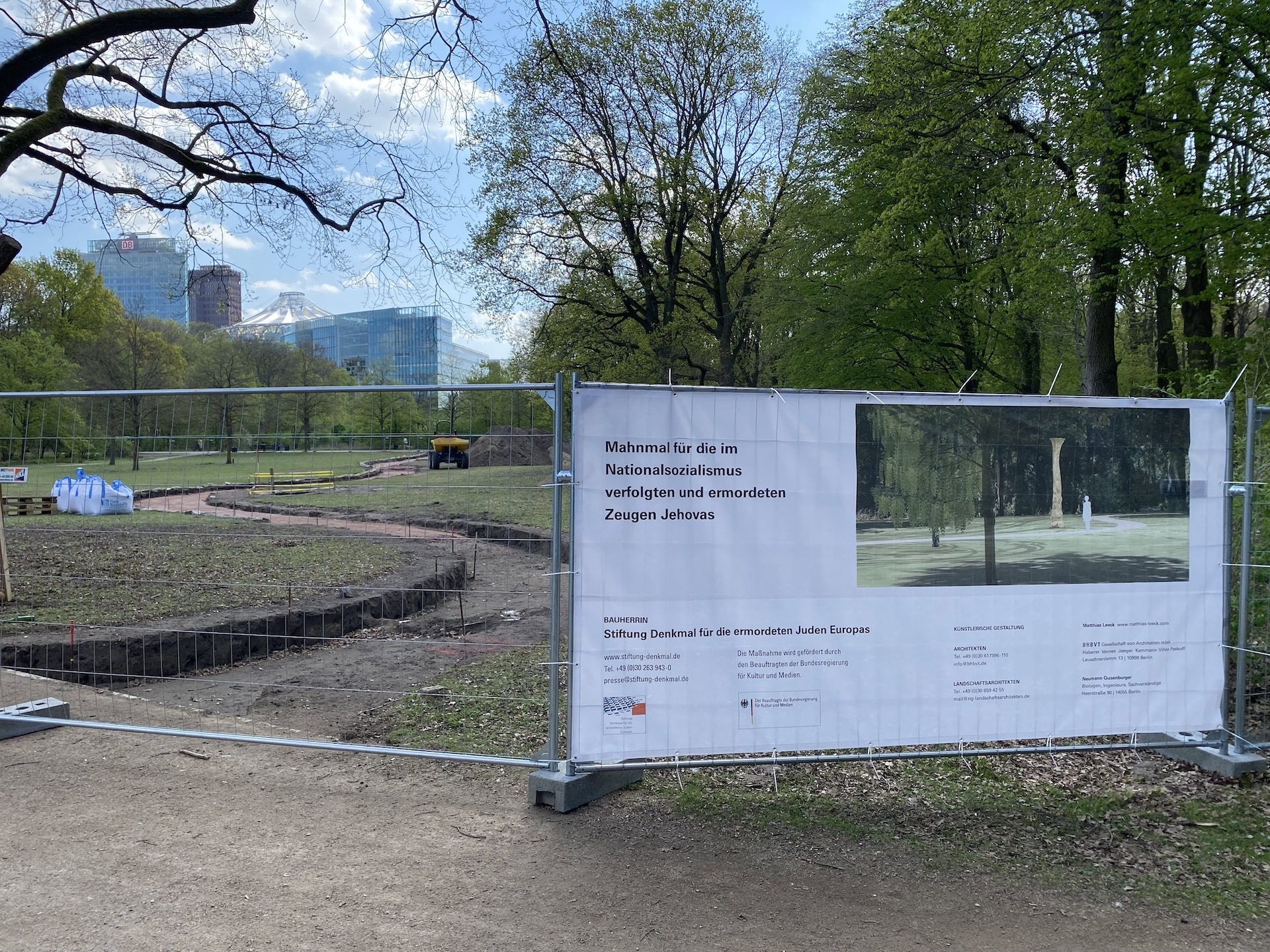 Gesamtansicht der Baustelle des Mahnmals für die im Nationalsozialismus verfolgten und ermordeten Zeugen Jehovas im Berliner Tiergarten. Im Vordergrund das Baustellenbanner am Bauzaun, dahinter die geöffnete Fläche mit Erdarbeiten und Baufahrzeugen. Am Horizont zeichnen sich die Gebäude am Potsdamer Platz ab. Rechts im Hintergrund ist der Baumbestand des Tiergartens zu sehen.