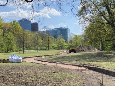 Blick über die Baustelle des Mahnmals im Berliner Tiergarten. Der geschwungene Zuweg aus rotbraunem Kies führt durch eine Grünfläche zum Baustellenbereich. Im Hintergrund sind die Hochhäuser am Potsdamer Platz und das Zeltdach des Sony Centers zu erkennen. Links im Vordergrund stehen Baumaterialien auf Paletten.