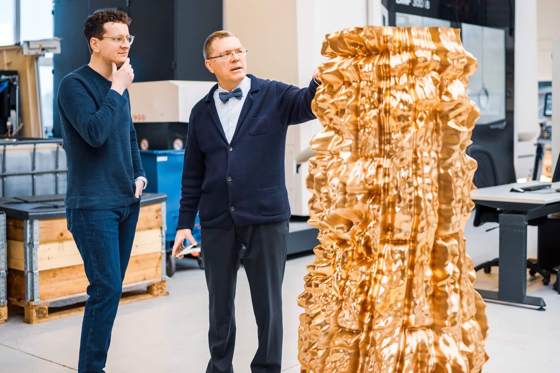Künstler Matthias Leeck und Uwe Langhals von der Arnold-Liebster-Stiftung betrachten ein Segment des Mahnmals für die verfolgten Zeugen Jehovas in einem Metallbetrieb in Wermelskirchen. Die bronzefarbene Skulptur zeigt eine stark zerklüftete Oberflächenstruktur.