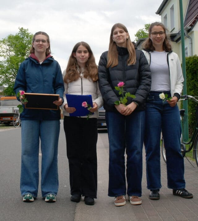 Vier Schülerinnen der AG »Kriegsgräber« der Europaschule Rövershagen stehen nebeneinander auf einem Gehweg in Ueckermünde. Sie tragen alltägliche Freizeitkleidung mit Jeans und Jacken in Blau-, Grau- und Weißtönen. In den Händen halten sie rosa und weiße Rosen, eine der Schülerinnen trägt zusätzlich ein Bild mit Holzrahmen, eine andere eine blaue Mappe mit der Biografie – Materialien für die Gedenkrede zur Stolpersteinverlegung für Martha Gehrke.