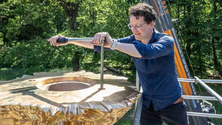 Matthias Leeck zieht auf einem Gerüst eine Gewindestange im Zentrum einer bronzenen Ronde an; im Hintergrund ein Kran und Bäume.