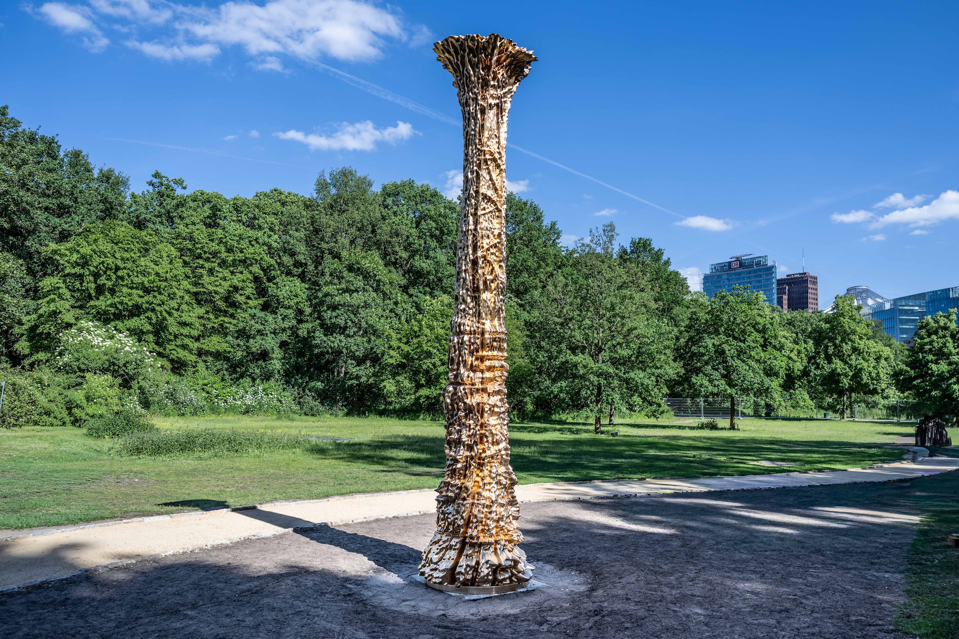 Das fertige Mahnmal in Form eines hohen, goldglänzenden Baumstamms steht auf einer Lichtung im Berliner Tiergarten; im Hintergrund Bäume und die Hochhäuser am Potsdamer Platz.