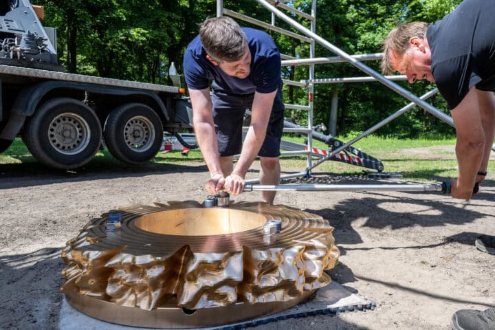 Axel Selbach und Florian Remscheid ziehen mit einer langen Stange eine Verschraubung an der untersten, auf dem Betonfundament liegenden Bronzeronde fest.