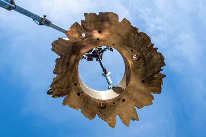 Untersicht einer einzelnen bronzenen Ronde, die an einem Kran vor blauem Himmel schwebt; im hohlen Zentrum sind Löcher für Gewindestange und Verschraubung zu erkennen.